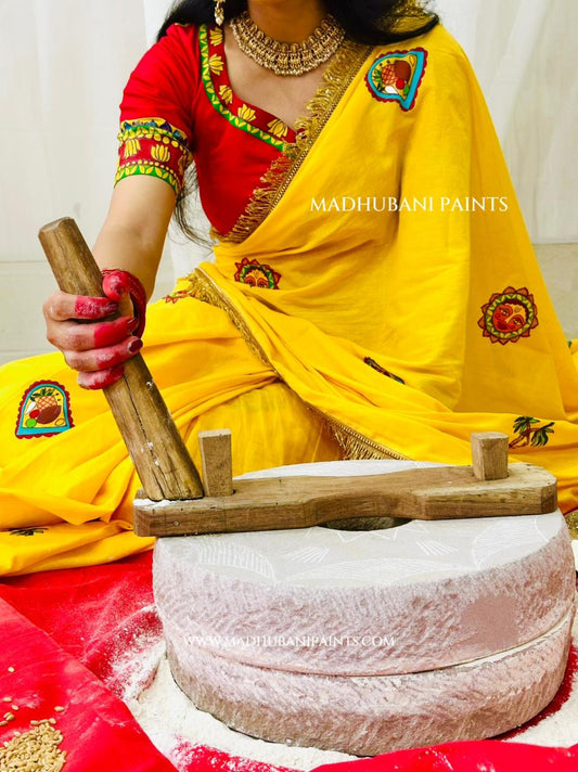 A woman in a bright yellow and red handpainted sari uses a wooden pestle to grind white powder in a stone mill for chhath puja.