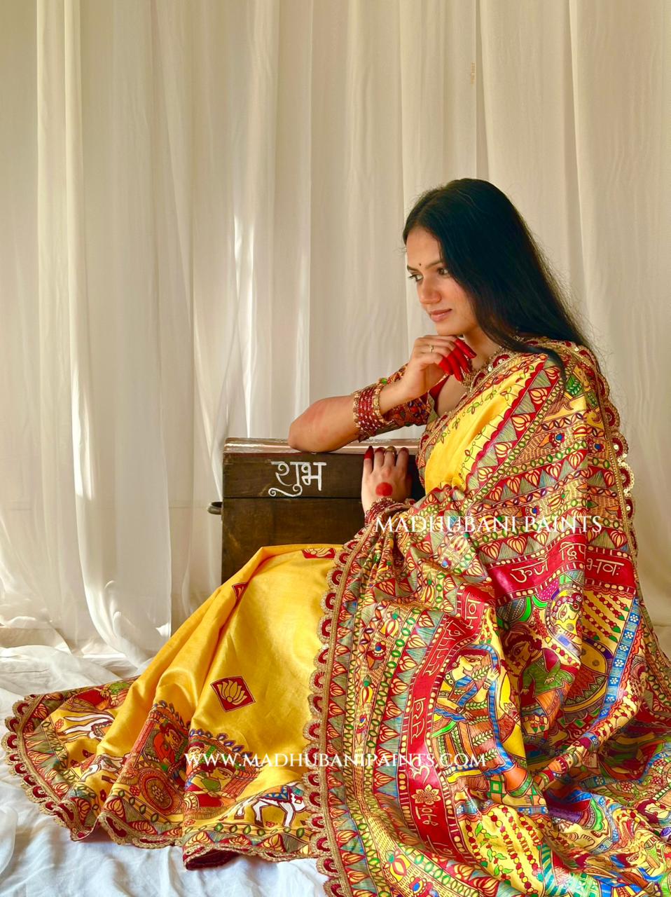 A woman wearing a traditional Indian saree sits on a bed in front of white curtains. The saree is bright yellow with an ornate multicolor border featuring red, green, blue, and gold patterns.