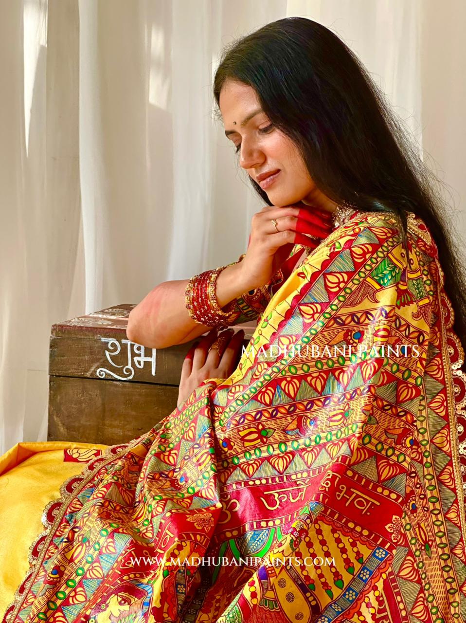A woman wearing a vibrant, multicolored traditional Indian saree with intricate hand-painted patterns in red, yellow, green, and gold. She is posed next to a rustic wooden box in front of a soft white curtain background.