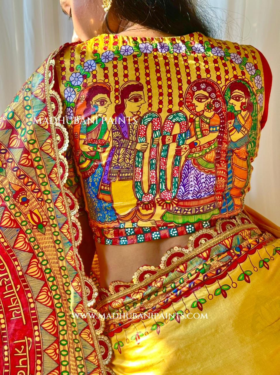 A close-up back view of a bride wearing a richly decorated traditional Indian blouse and saree. The blouse features vibrant hand-painted Madhubani-style figures and patterns in red, green, blue, purple, and yellow on a golden-yellow base.