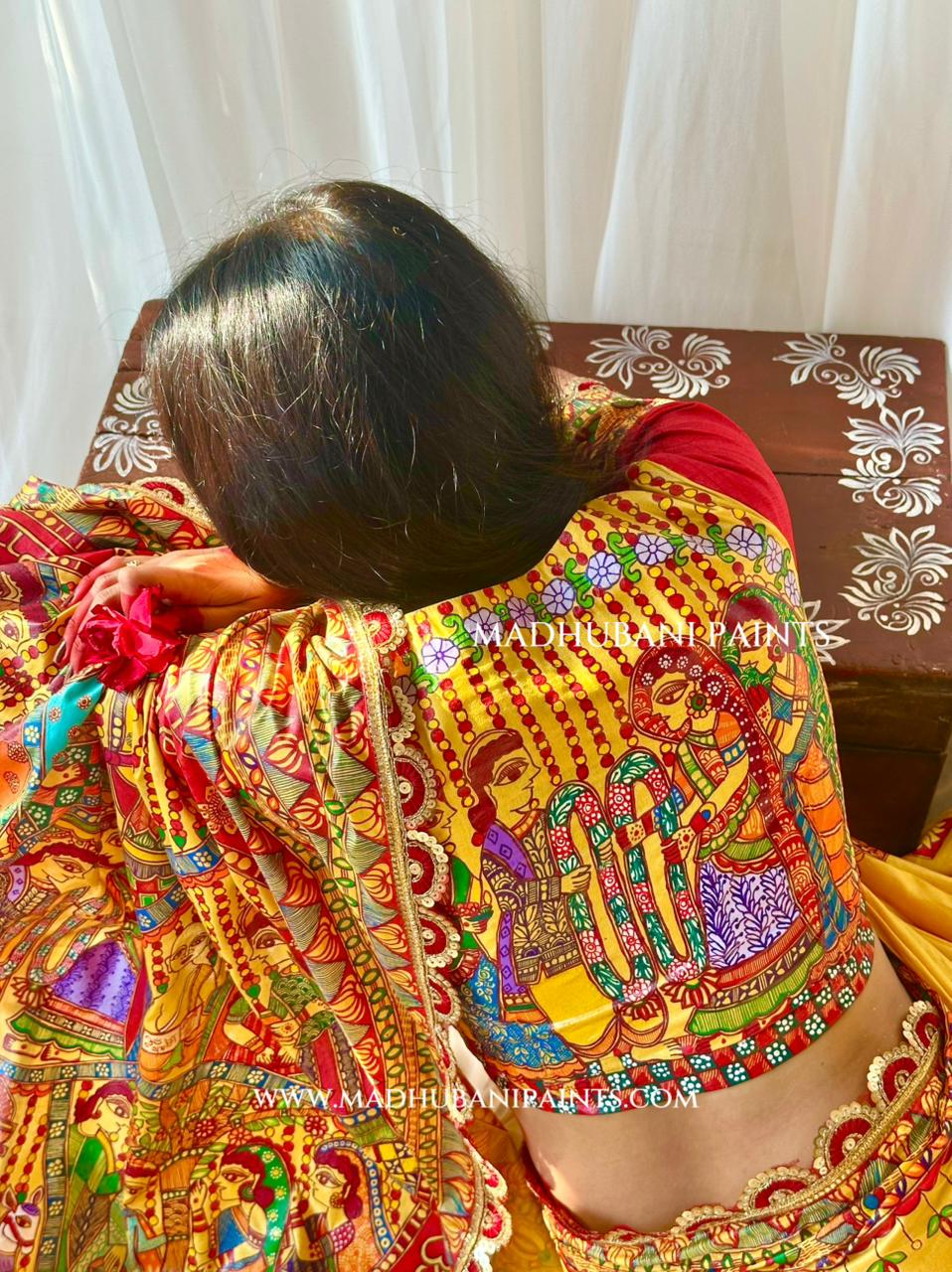 A woman viewed from behind wearing a highly detailed, colorful traditional Indian blouse with intricate hand-painted motifs, set against a background of white curtains and a wooden table.