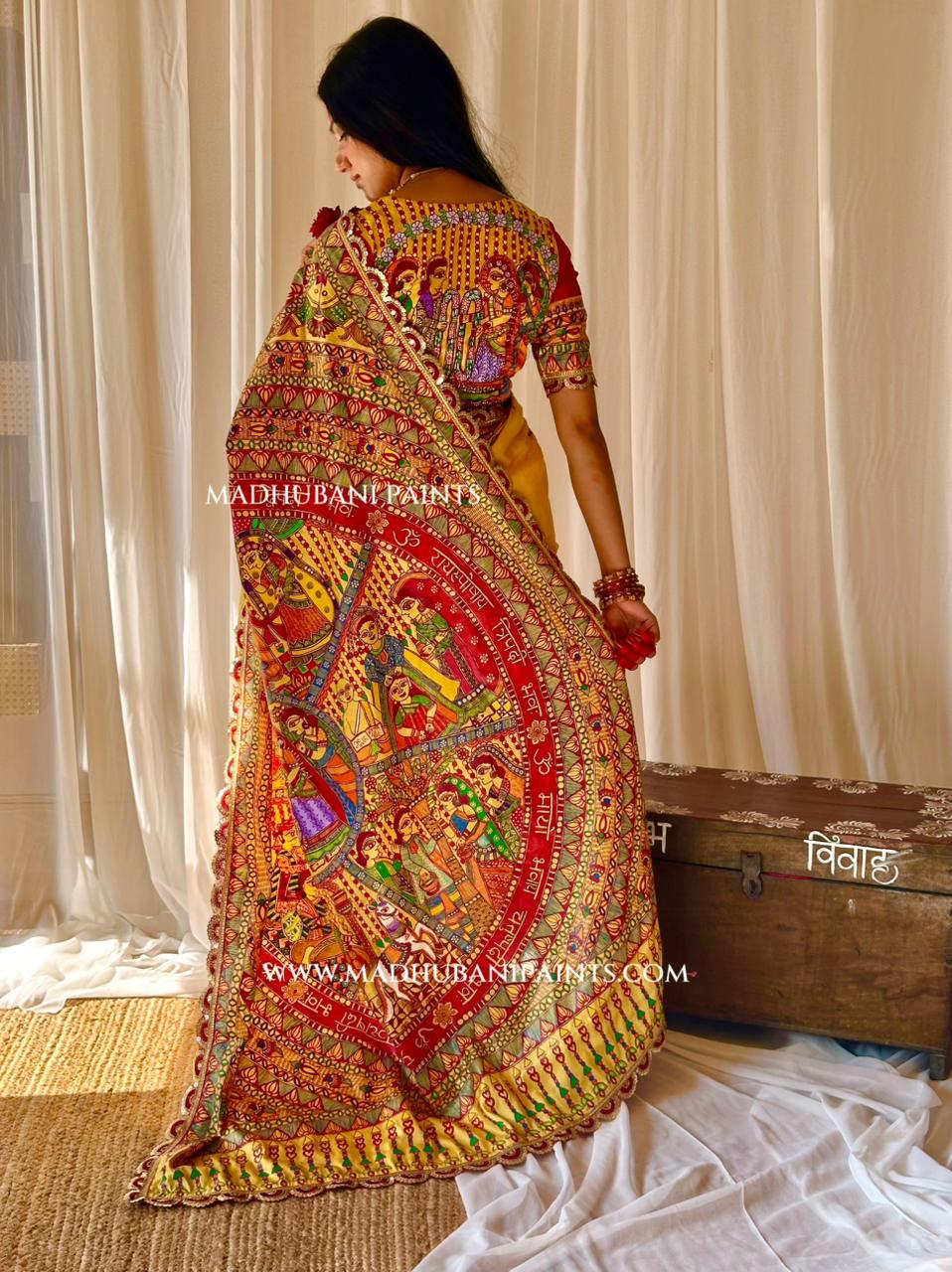 A woman wearing a highly detailed, multicolored Indian madhubani painting saree with intricate hand-painted style motifs, standing in front of cream-colored curtains and next to a rustic wooden chest.