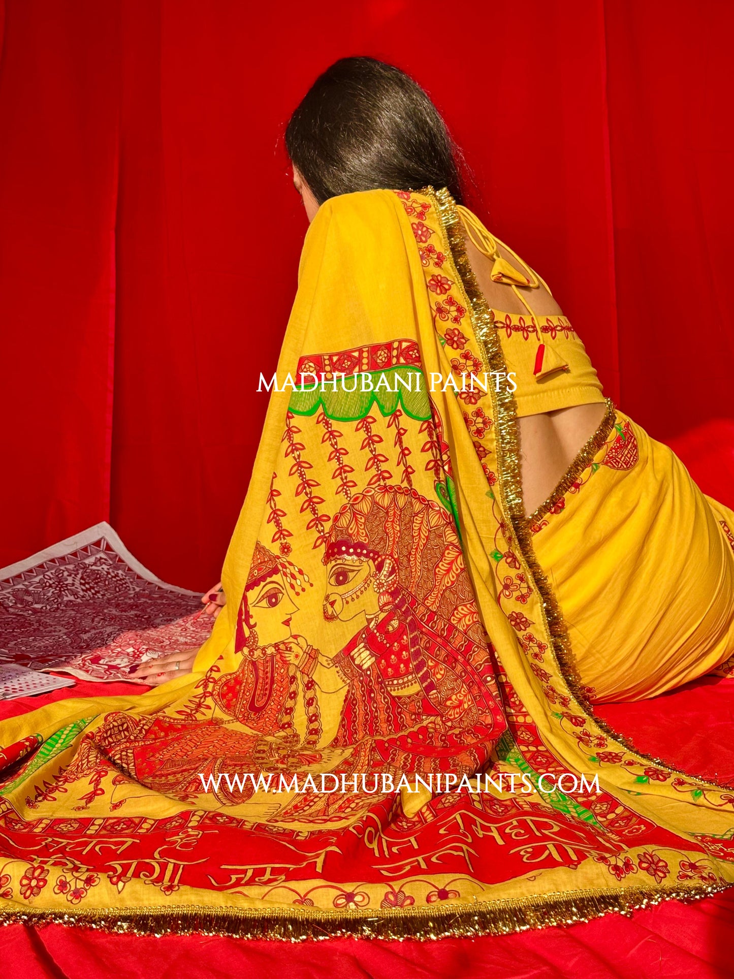 Woman wearing a yellow saree with intricate red and green designs on a red background, featuring Madhubani Paints branding.