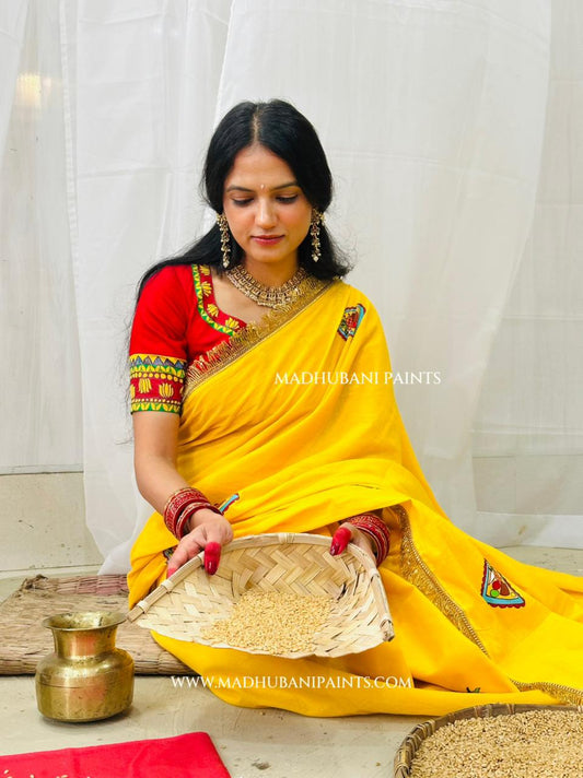 A woman in a chhath puja bright yellow saree with a red hand-painted blouse sits on the floor holding a woven tray of grains, with a brass pot and more grains on the floor in front of a white curtain backdrop.
