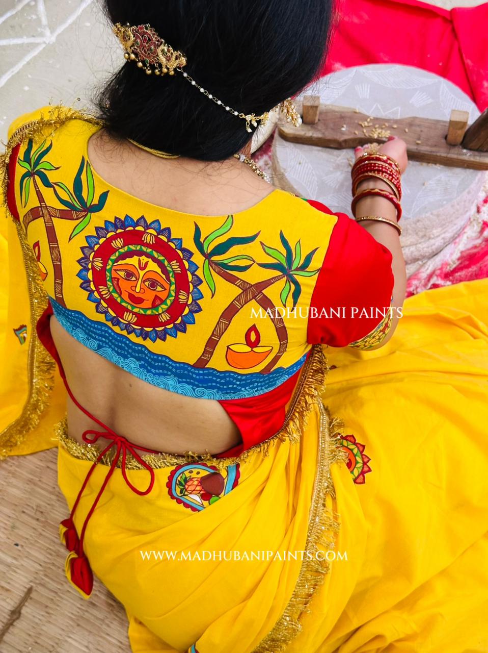 A woman viewed from behind wearing a bright yellow chhath puja saree with a red sleeve and a heavily madhubani painting back panel featuring a colorful sun motif, palm trees, and a diya. 