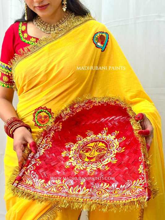 A woman wearing a bright yellow chhath puja sari holds a large red decorative tray with gold and yellow painted motifs. The sari has golden lace details