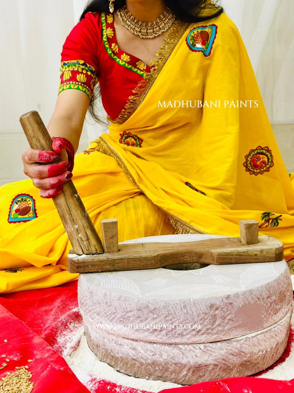 A woman in a bright yellow and red handpainted sari uses a wooden pestle to grind white powder in a stone mill for chhath puja.
