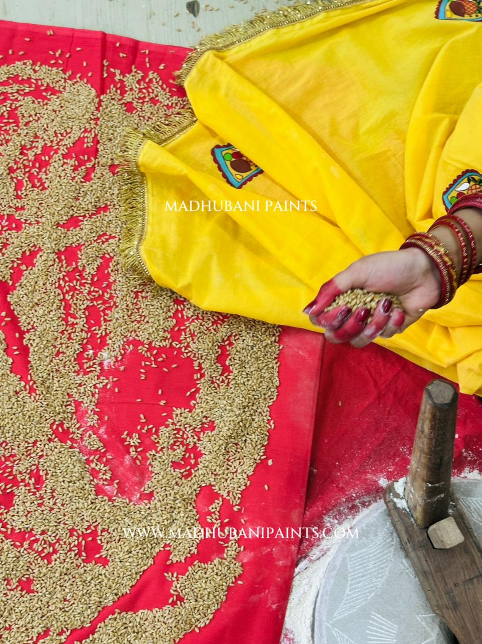 A close-up of a traditional Indian textile setup showing a bright yellow fabric with gold border details and a red fabric spread underneath, both covered with scattered beige grains. A person's hand with red nail polish and bangles is holding some of the grains.