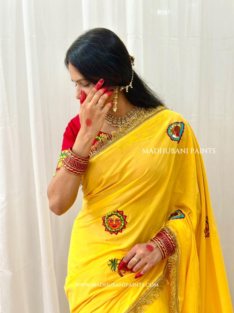 A woman wearing a bright yellow  chhath puja madhubani painting saree with stitched blouse set in red and gold embroidery, a red blouse, and traditional jewelry, standing in front of a white curtain. 