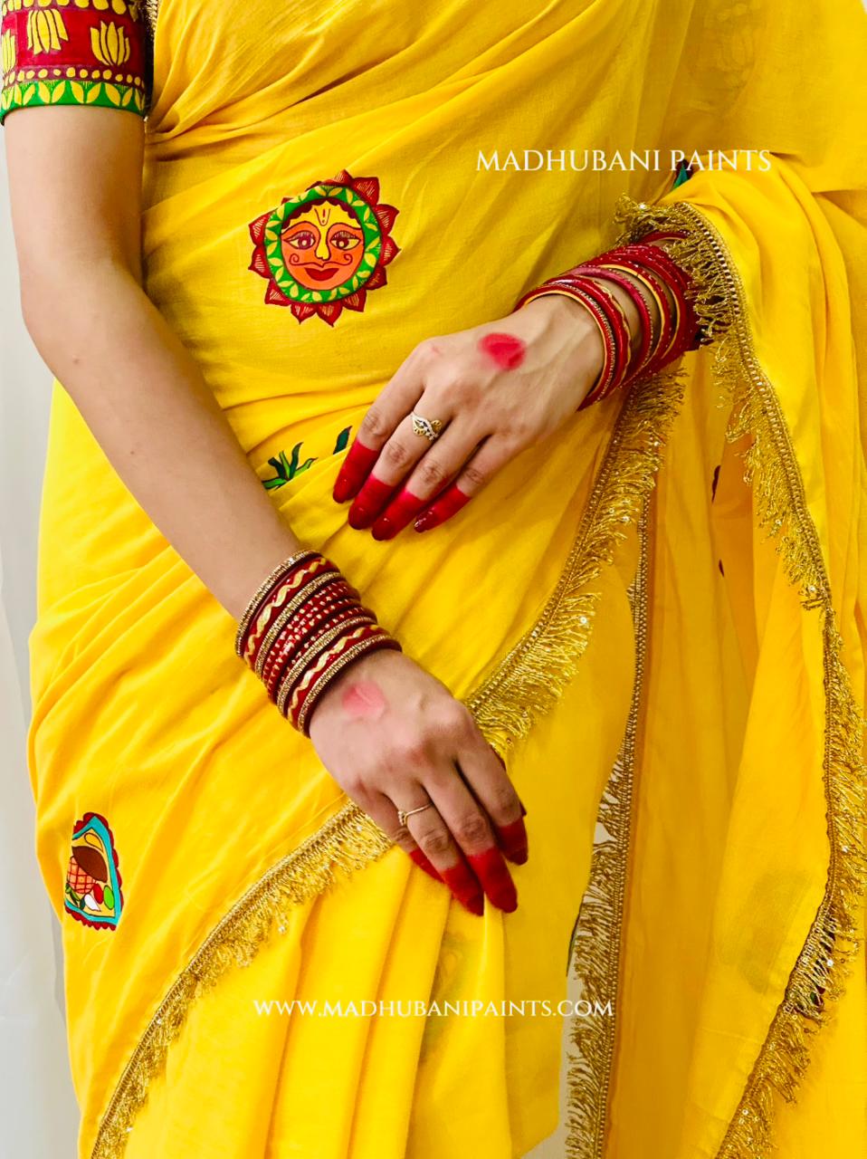 A close-up of a person wearing a bright chhath puja yellow sari with suraj motifs, red bangles, and red nail polish. The sari has gold fringe along the border and a matching blouse with colorful madhubani painting.