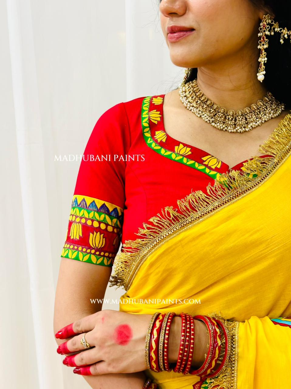A close-up of a woman wearing a bright yellow and red traditional madhubani painting Indian saree with colorful handpainted borders and gold zari detailing. She is also wearing a heavy gold necklace, earrings, and red bangles.