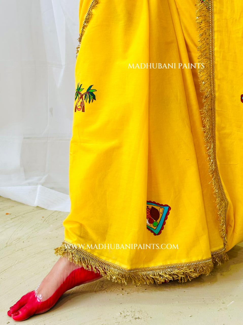 A close-up of a pleats of a bright yellow chhath puja saree, with gold fringe trim and small surya madhubani painting motifs, shown on a model wearing a red pointed shoe.