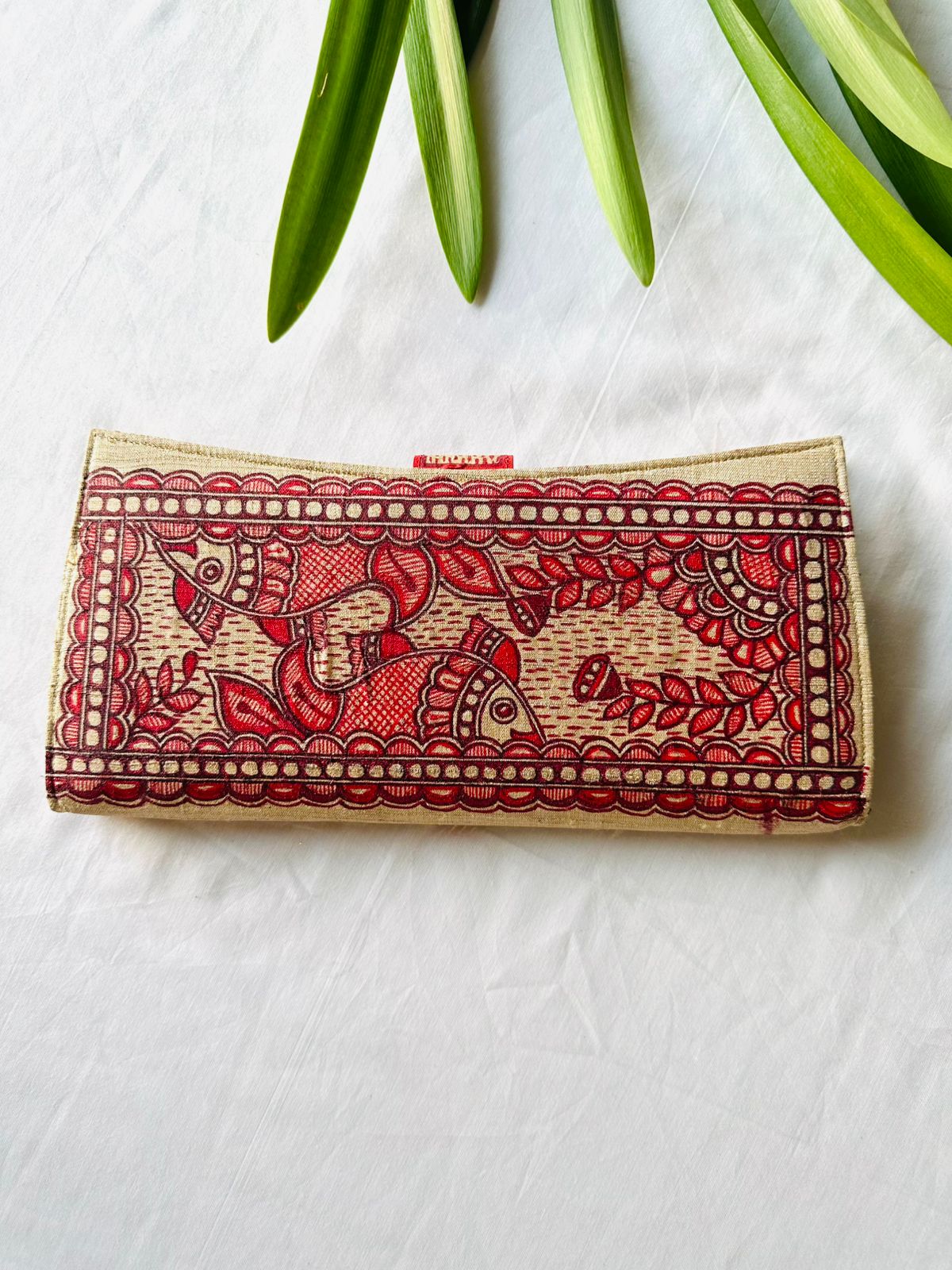 A rectangular clutch bag with a beige base and intricate red and black hand-painted batik-style elephant and floral patterns, styled on a white fabric background with green leaves at the top.