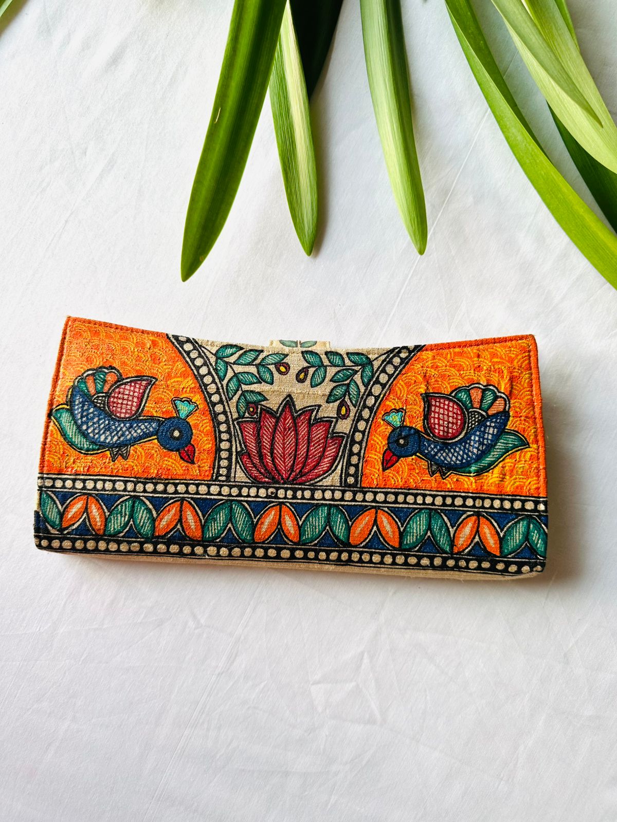 Hand-painted Madhubani clutch in vivid orange with two birds facing a central lotus flower beneath a leafy arch