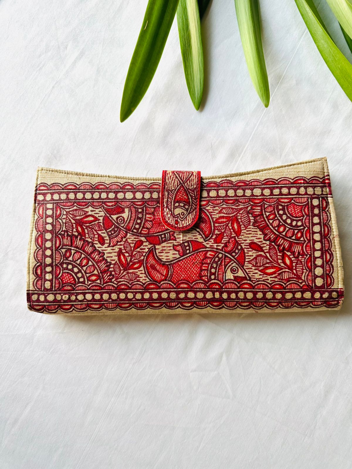 A rectangular clutch bag with an intricate red and beige hand-painted folk art pattern, featuring a central flap closure, laid flat on a white fabric background with green leaves at the top.