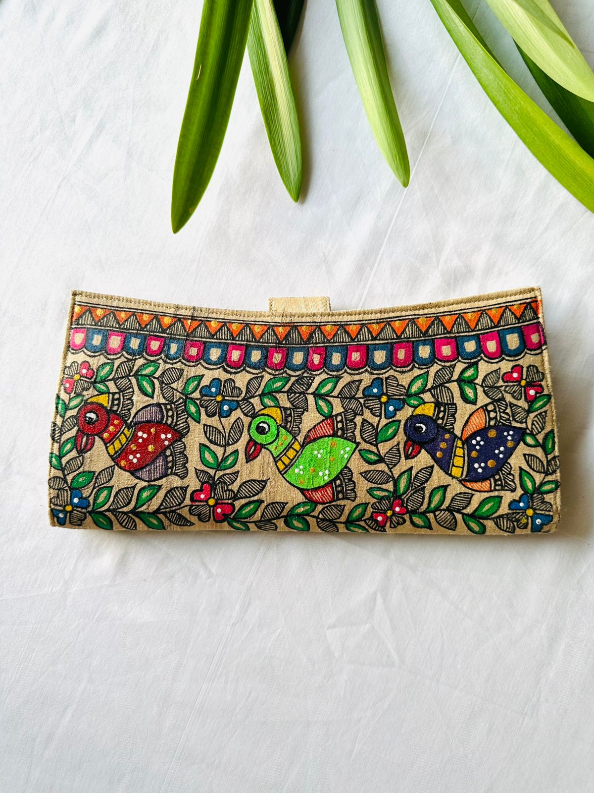 Hand-painted Madhubani clutch with three colorful birds in red, green, and navy perched among leaves and flowers, framed by a pink and blue triangle border