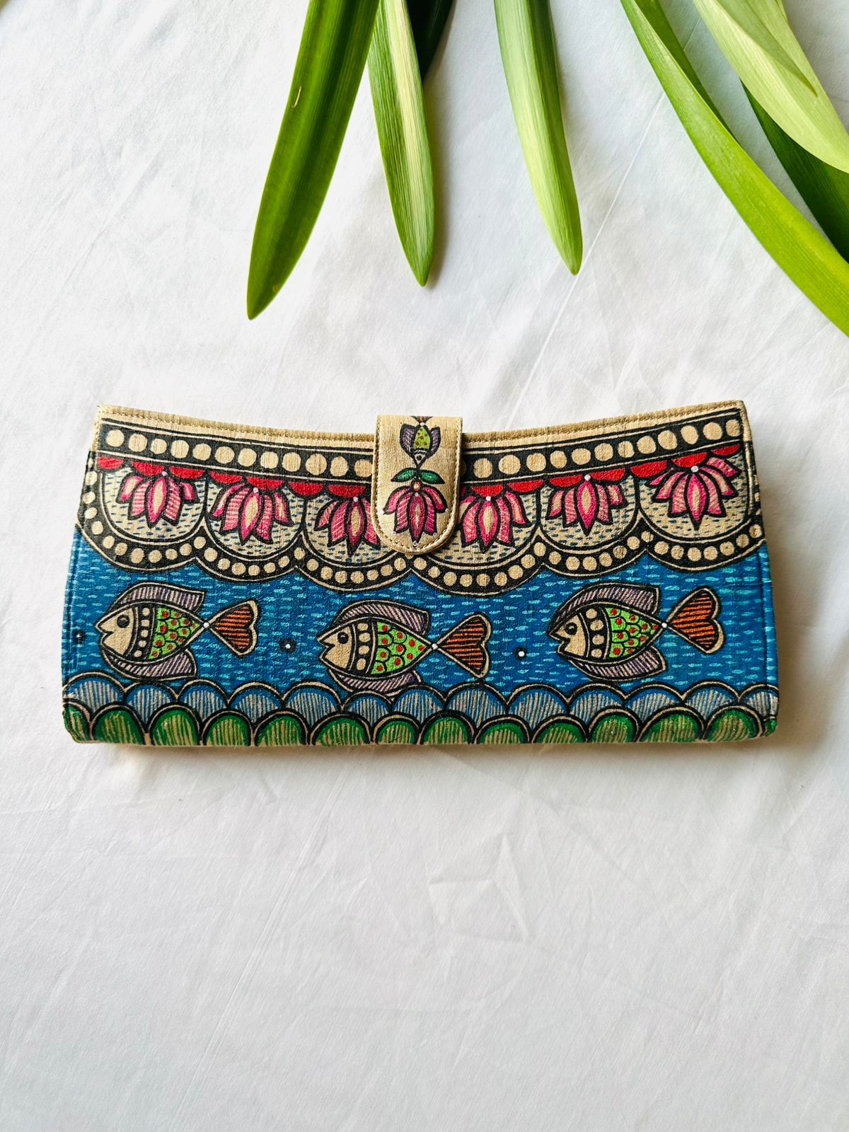 Hand-painted Madhubani clutch in blue with three fish swimming, a scalloped pink lotus border above and a green scaled border below, a lotus on the tab closure