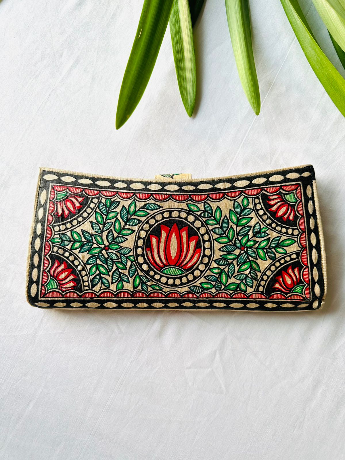 Hand-painted Madhubani clutch in cream with a red lotus in a round medallion at the center, flanked by green leaf sprays and small lotus flowers in each corner.