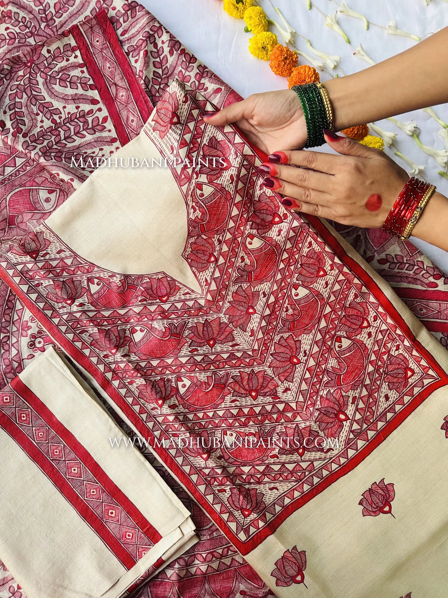 A woman wearing green and red bangles spreads out a cream suit fabric hand-painted with red Madhubani fish and lotus designs.