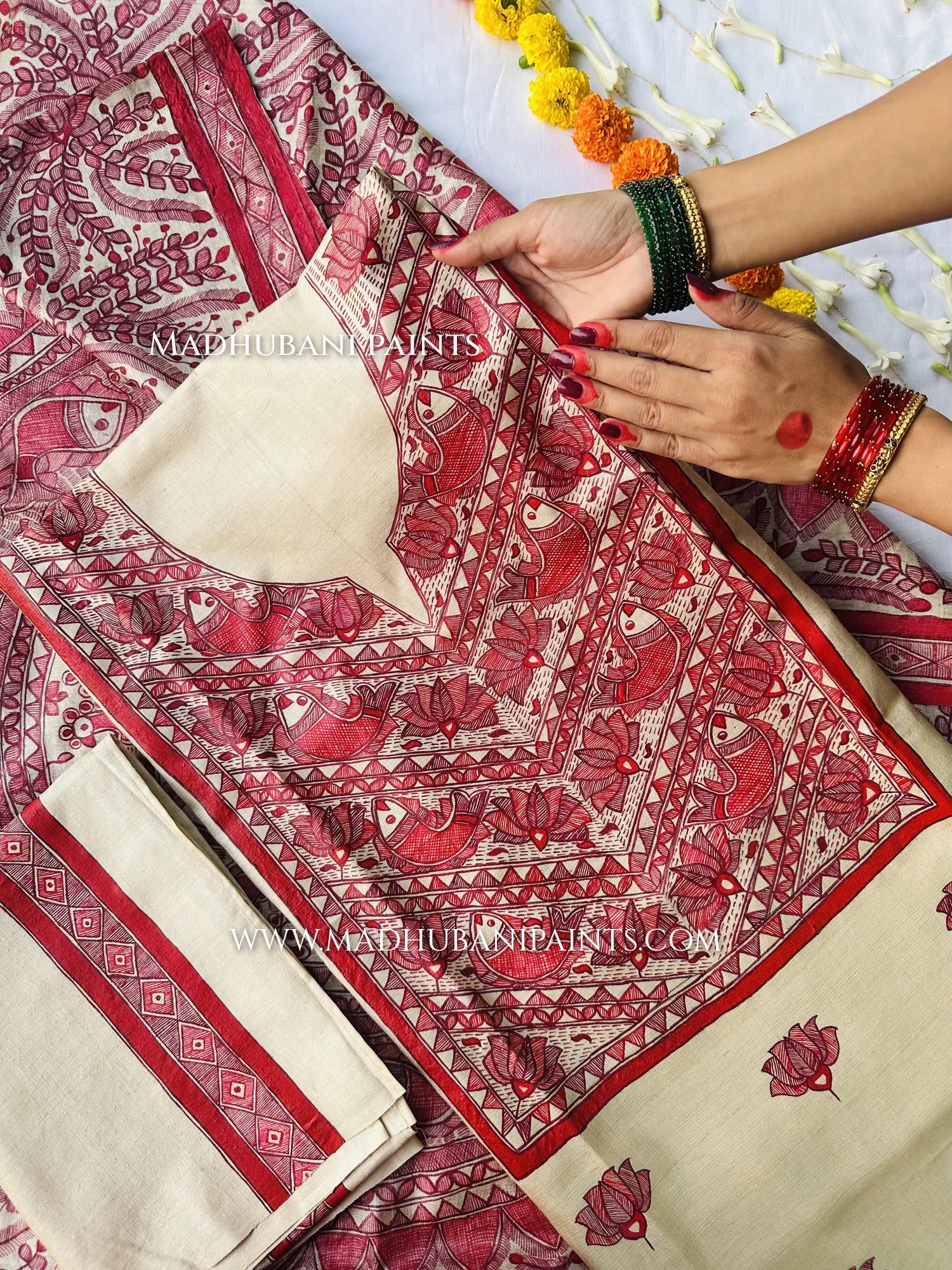 A woman wearing green and red bangles spreads out a cream suit fabric hand-painted with red Madhubani fish and lotus designs.