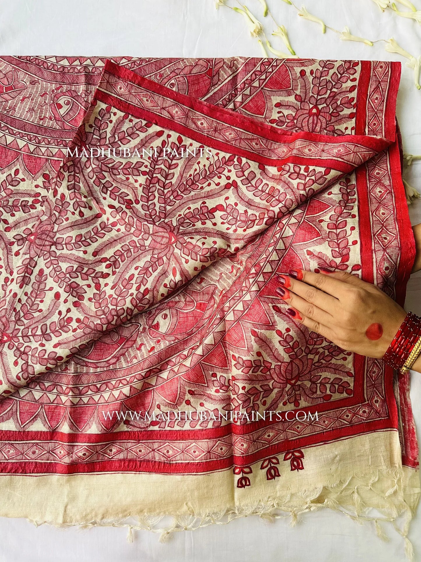 A woman in green bangles holds a draped cream dupatta hand-painted with red Madhubani leaf and vine patterns.