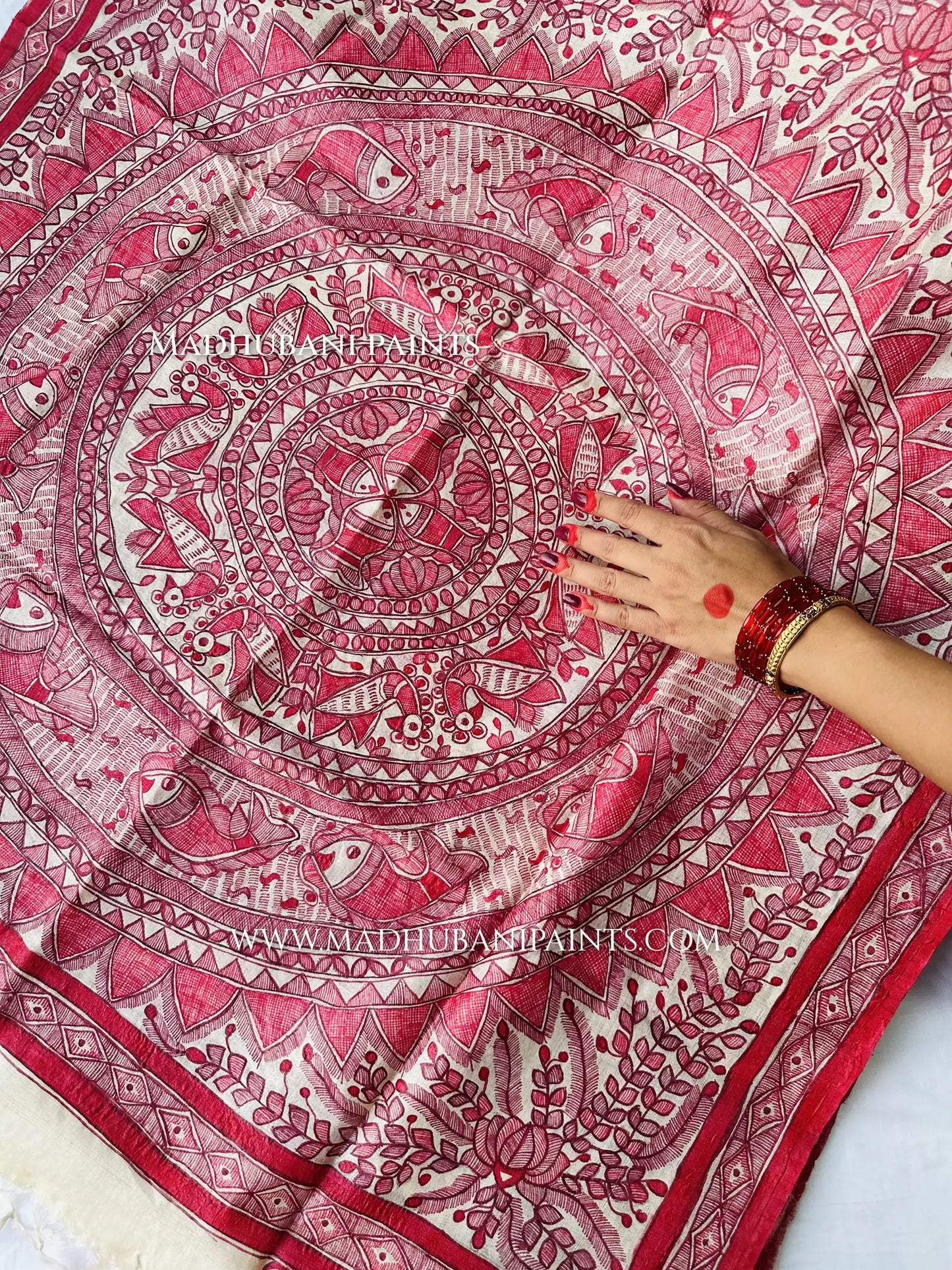 A hand rests on a cream dupatta covered in a detailed red Madhubani mandala with fish, birds and lotus motifs.