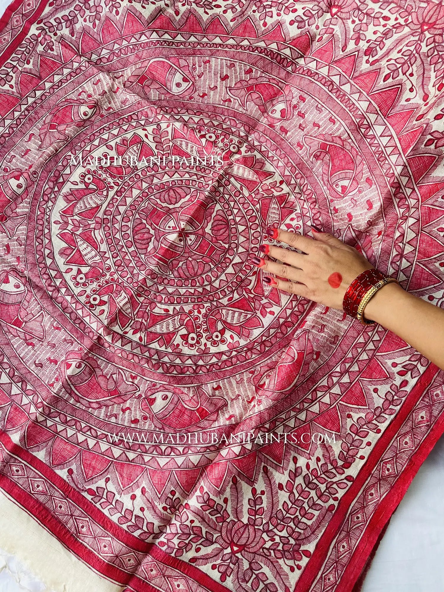 A hand rests on a cream dupatta covered in a detailed red Madhubani mandala with fish, birds and lotus motifs.