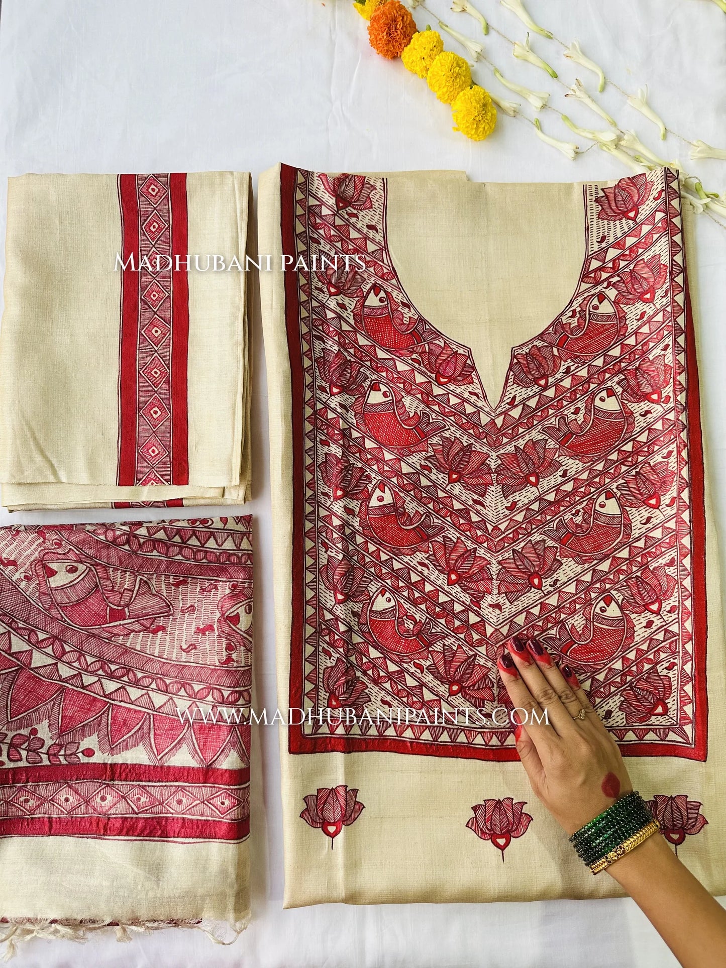 Hand-painted Madhubani unstitched suit in cream and red, with a painted neck yoke full of fish and lotus motifs, laid out with marigold flowers.