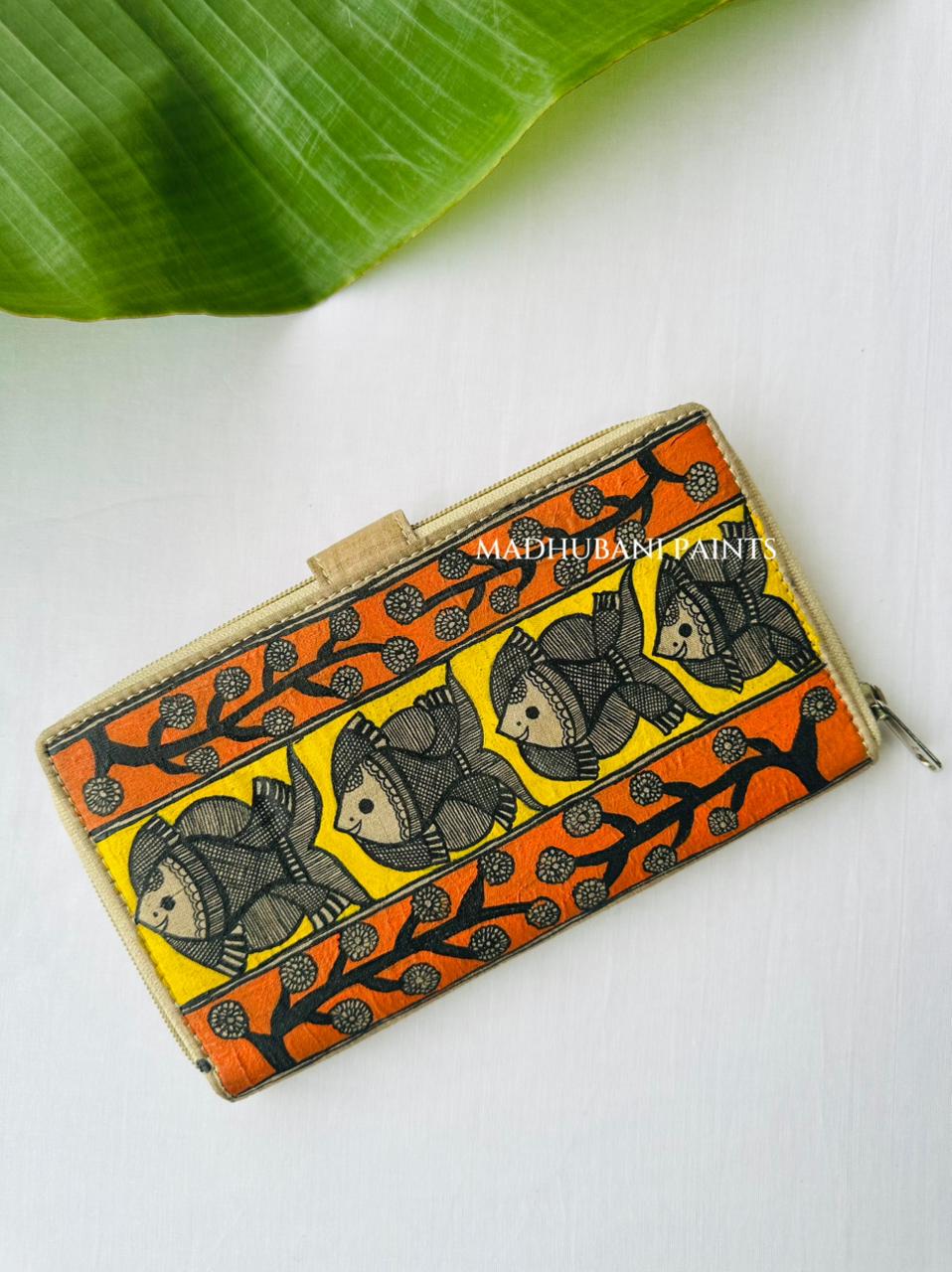 Hand-painted Madhubani clutch in yellow with black-and-cream fish scattered across the front, an orange border, and a fish painted on the tab closure.