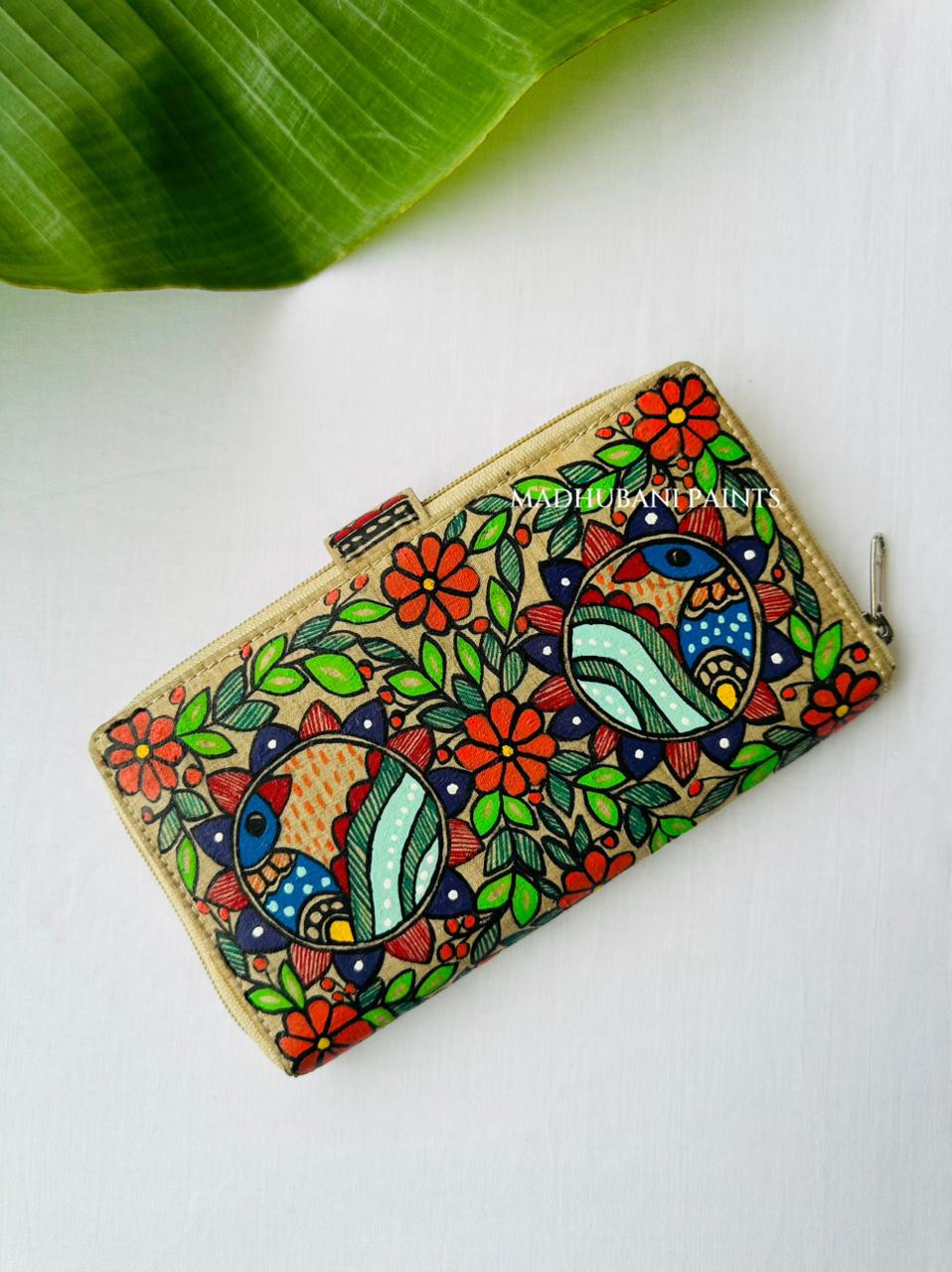 Back side of the Hand-painted Madhubani clutch in beige with two circular medallions of stylized peacocks in teal, red, and blue, surrounded by orange flowers and green leaves, a small fish painted on the tab closure