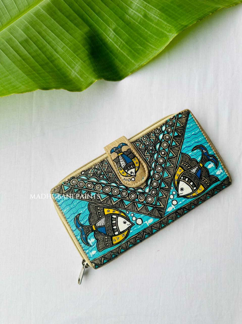 Hand-painted Madhubani clutch in teal with two fish facing each other over a wave pattern, a small fish painted on the tab closure, and a triangle-and-dot border