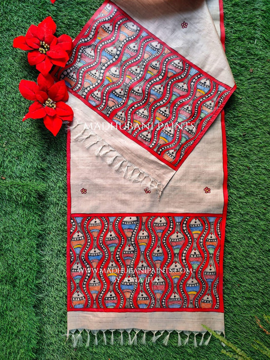 A red and beige hand-painted Madhubani Tussar silk stole with fish motifs displayed on a grassy surface, accompanied by red flowers.