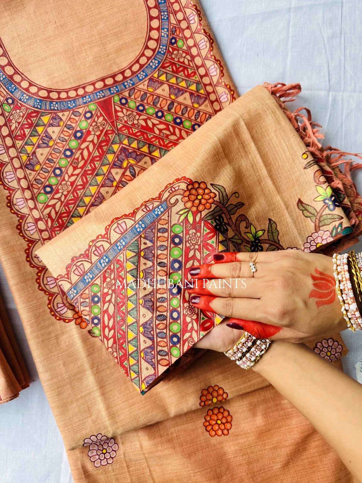 A close-up of a beige fabric textile with colorful painted geometric and floral patterns, draped over a matching folded piece. 