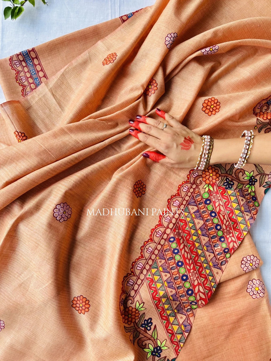 A peach-colored cotton dupatta with colorful geometric and floral motifs, draped elegantly with a hand resting on it. The fabric features a highly detailed, scalloped painted border with red, orange, green, and blue patterns.