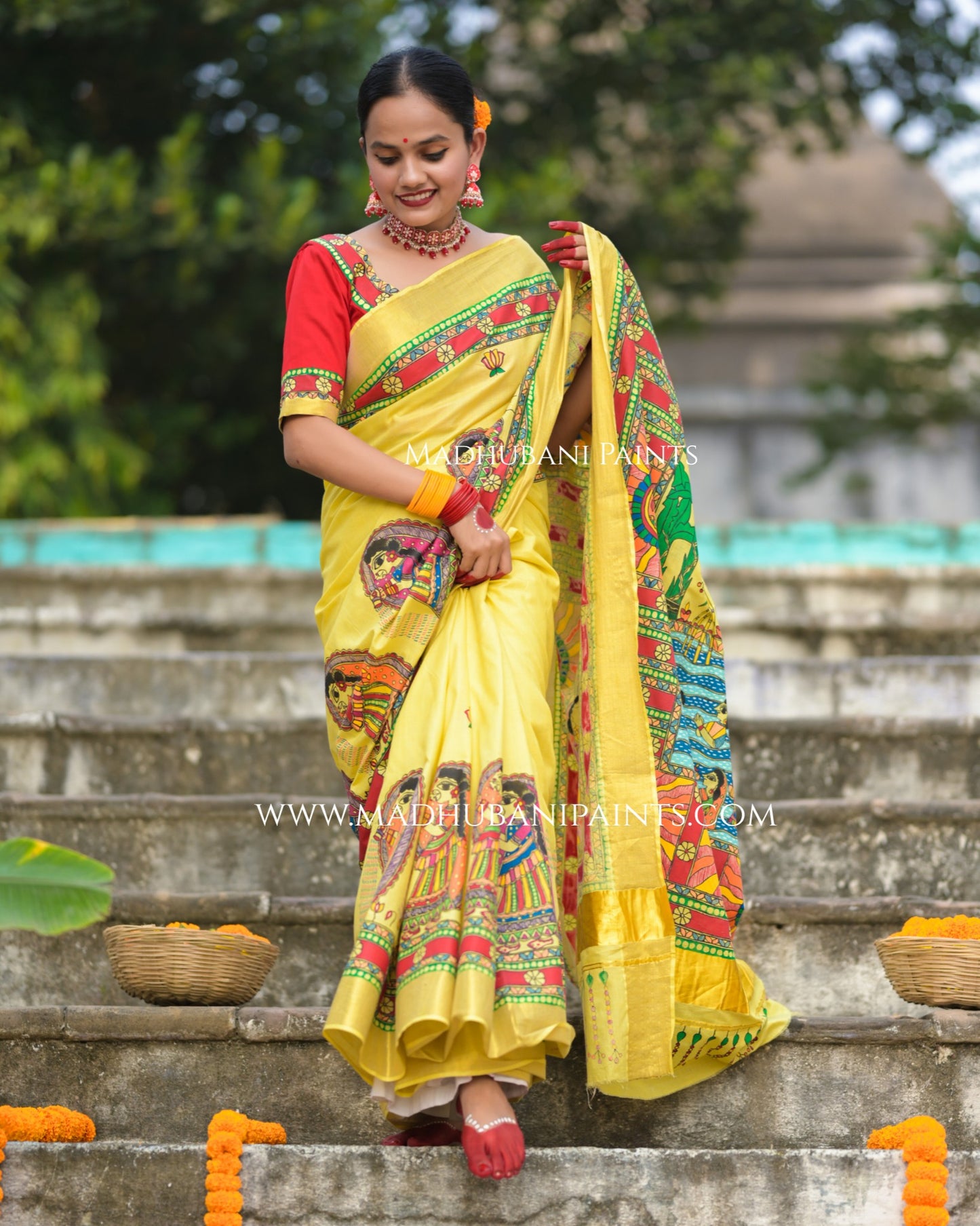 Woman in a yellow saree with red and green madhubani painting standing on steps outdoors.