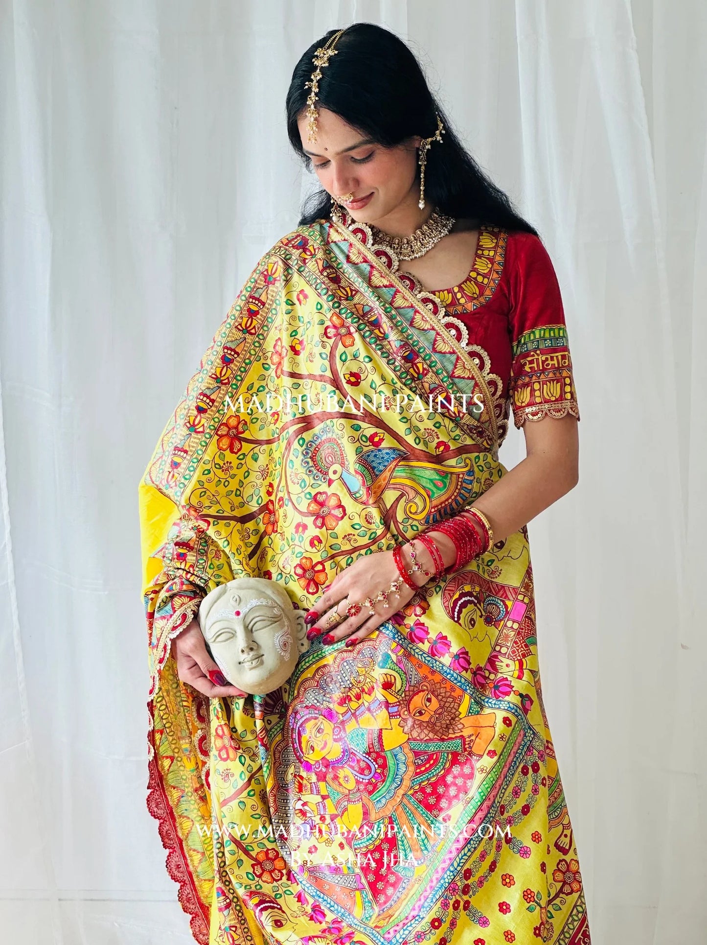 Model holding a traditional Devi face mask while wearing Devi Ki Sawari hand-painted Madhubani saree in festive yellow and red.