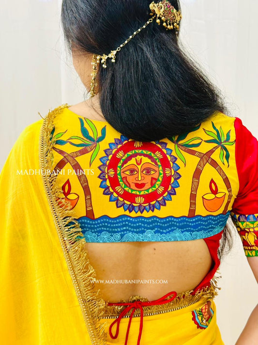 A woman viewed from behind wearing a bright yellow chhath puja saree with a red sleeve and a heavily madhubani painting back panel featuring a colorful sun motif, palm trees, and a diya. 