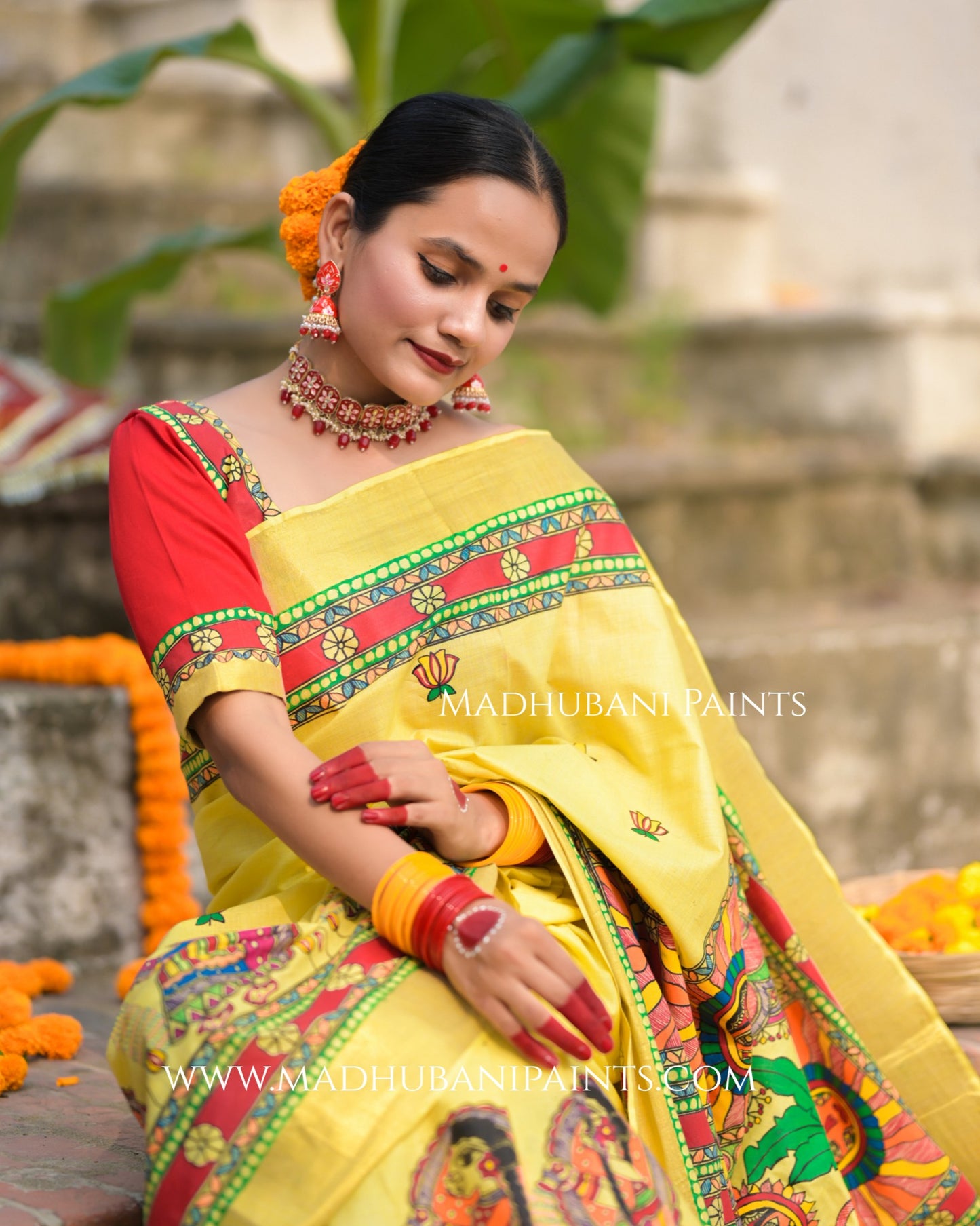 Woman in a yellow saree with red blouse, sitting outdoors with 'Madhubani Paints' branding.