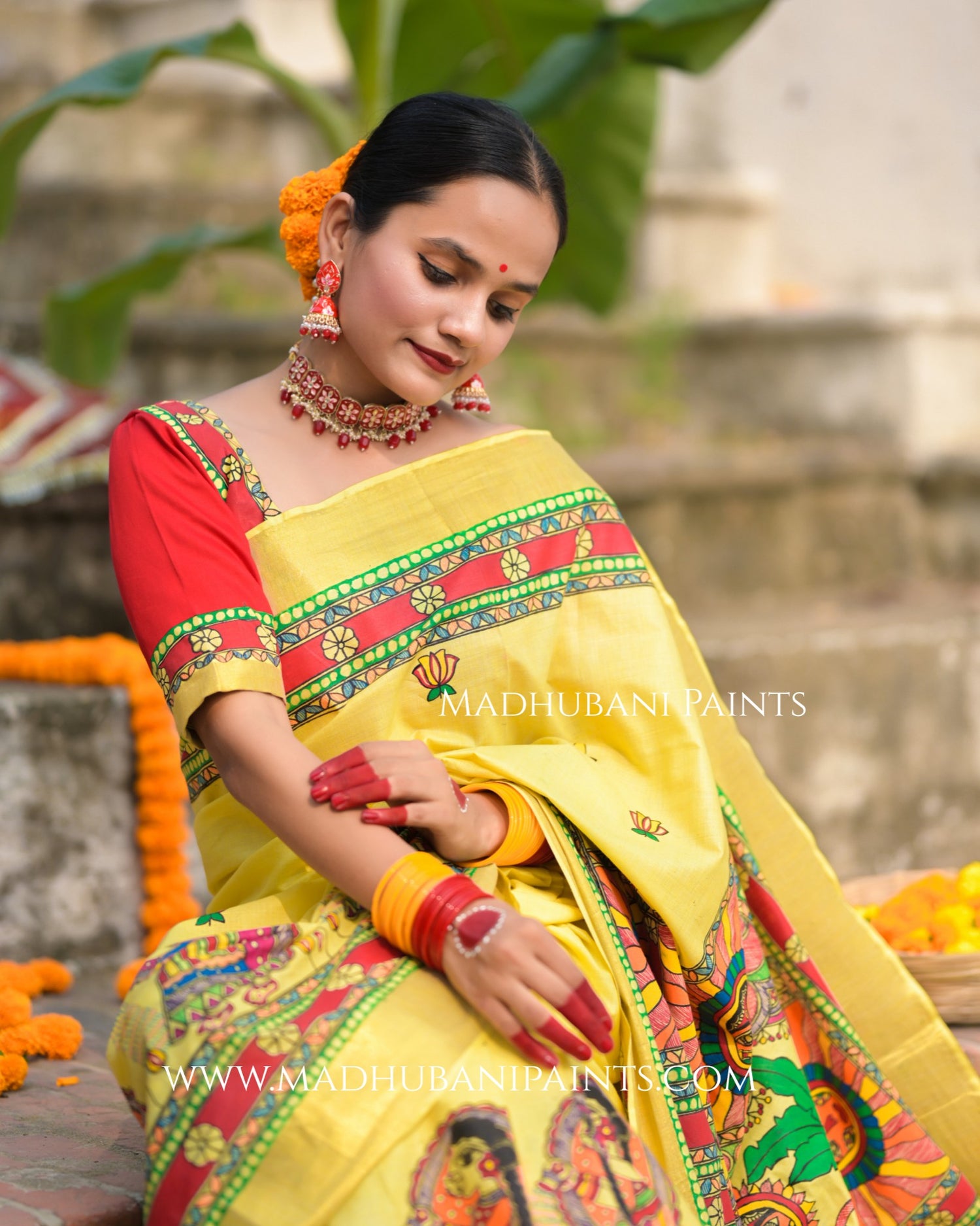 Woman in a yellow saree with red blouse, sitting outdoors with 'Madhubani Paints' branding.